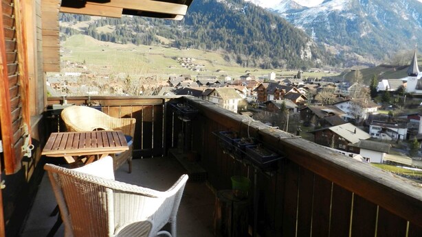 Een balkon met rieten stoelen, een houten tafel en uitzicht op een dorp en bergen.