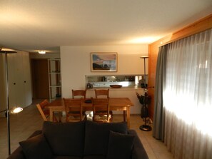 A living room with a sofa, a dining table with chairs, a kitchen area with a sink and cabinets, and a large window with curtains. - Hübeli (Chapman) Appartement/Fewo, Dusche oder Bad, WC, 2 Schlafräume by Interhome (Zweisimmen)