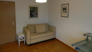 Een kamer met een beige bank, een klein wit bijzettafeltje, een houten deur en twee ingelijste foto's aan de muur.
