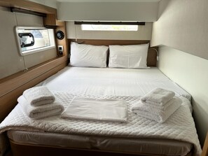 A neatly made bed with white linens and towels, a window with blinds, and a small control panel on the wall. - Sun-Kissed Sailing Adventure in Islas Sarónicas (Alimos)
