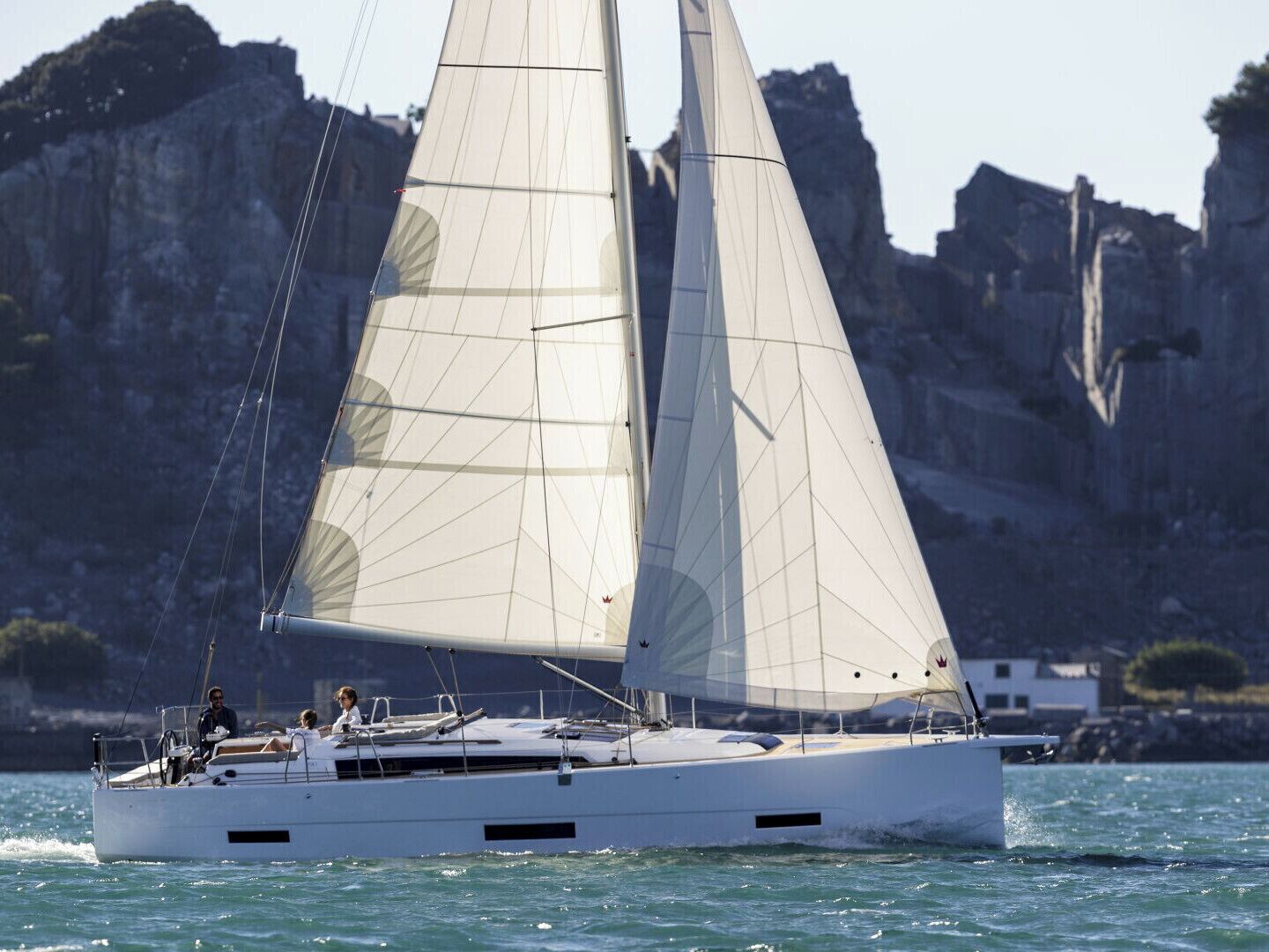 Un voilier à la coque et aux voiles blanches, naviguant sur des eaux calmes, avec des falaises rocheuses en arrière-plan.