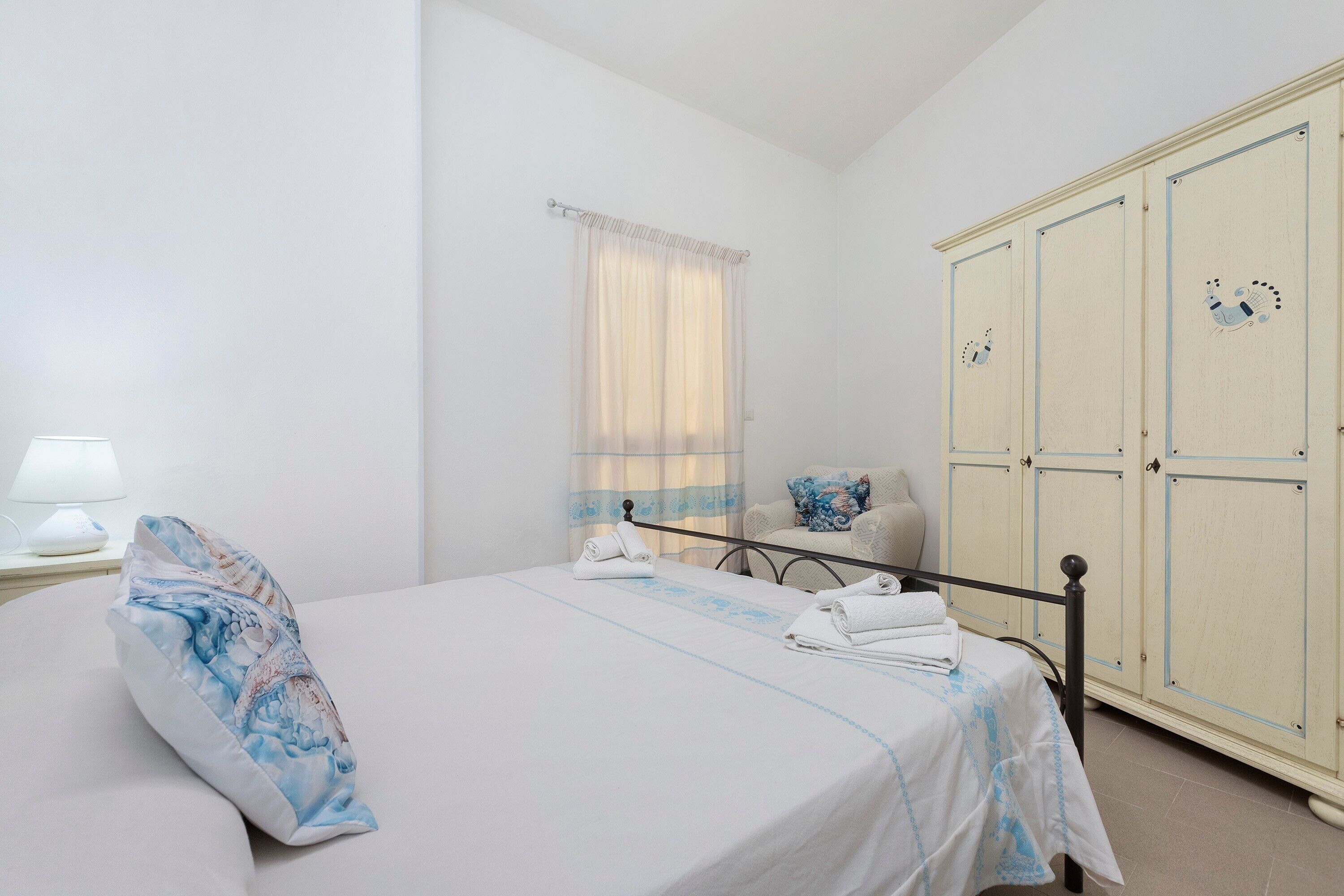 Quarto de hotel com cama, mesa de cabeceira, abajur, janela com cortinas, guarda-roupa e uma cadeira.
