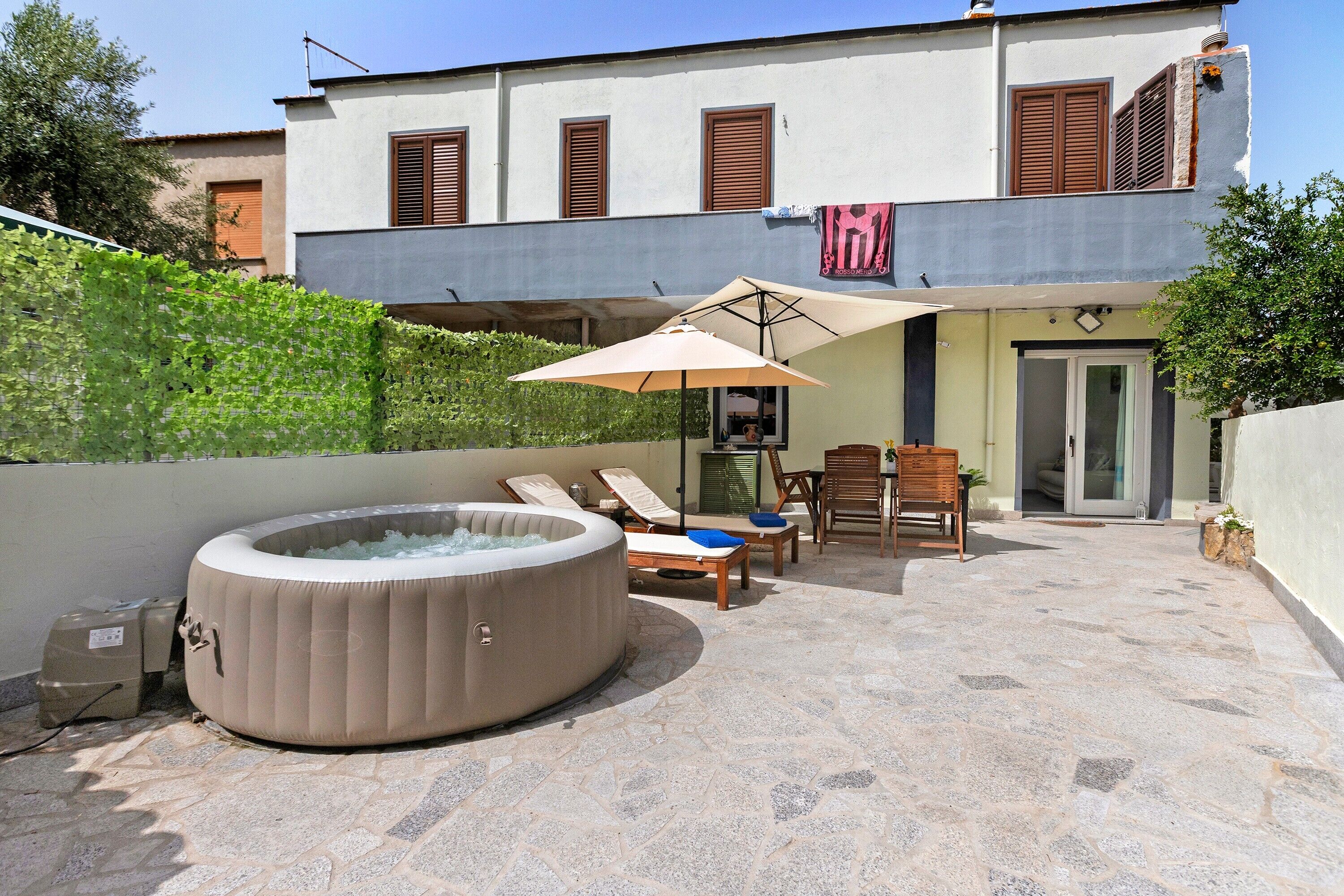 A hot tub with lounge chairs and an umbrella, a stone patio, and a building with shutters.