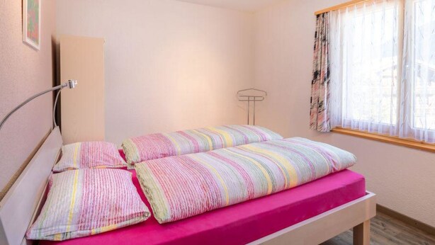 A single bed with a pink mattress and striped bedding in a room with a wooden floor and a window with sheer curtains. - Hübeli (Moosmatten) Appartement/Fewo, Dusche oder Bad, WC, 1 Schlafraum by Interhome (Zweisimmen)