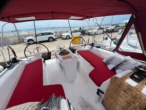 Schiffsinnenraum mit rotem Verdeck, weißen Kissen, einem kleinen Tisch und Blick auf das Meer sowie geparkte Autos.