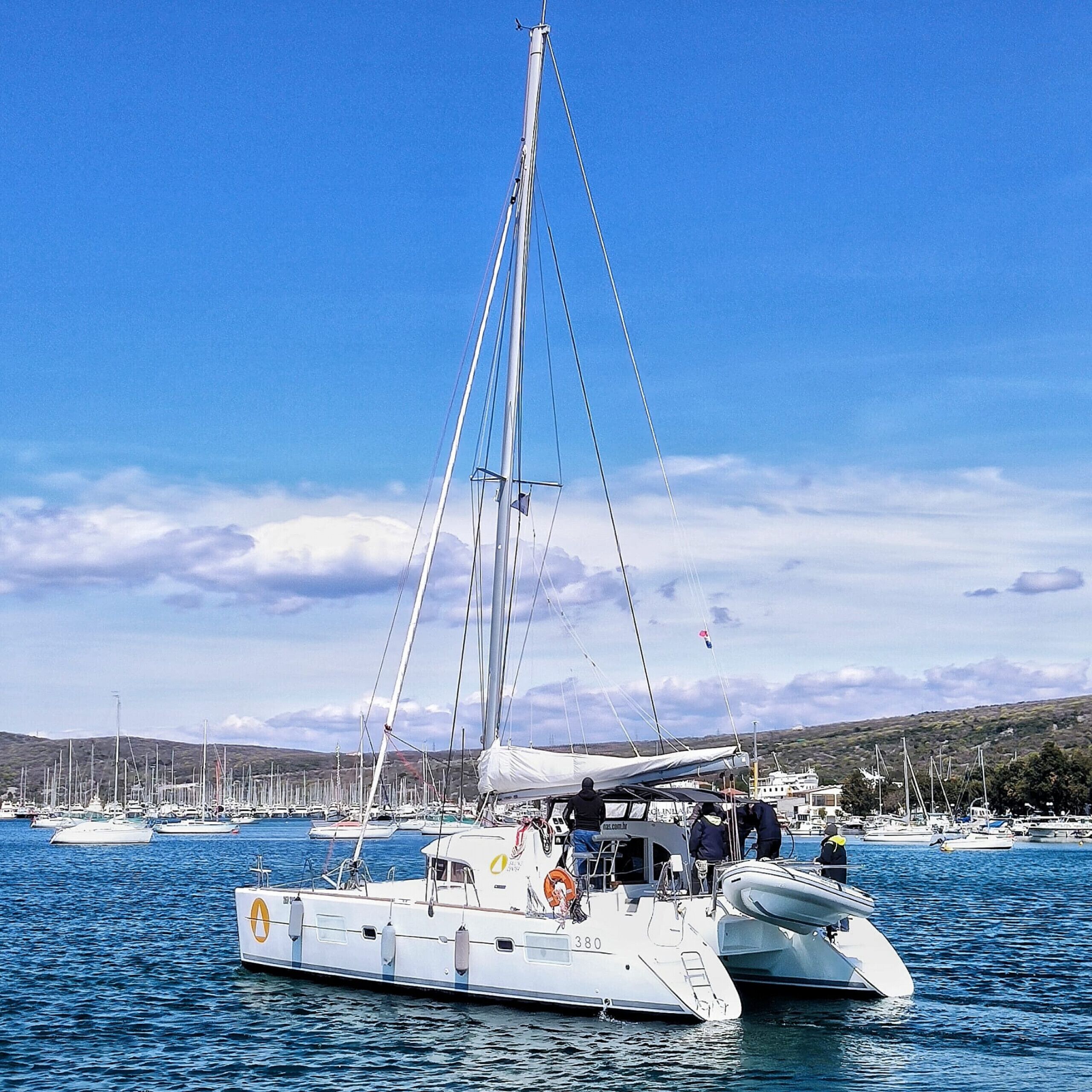 Un velero amarrado en un puerto deportivo con varios barcos, cielos despejados y un paisaje montañoso al fondo.