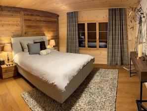 A bedroom with a large bed, wooden walls, a nightstand with a lamp, a dresser, and a window with curtains. - Heimersberg Appartement/Fewo, Bad, WC 3-Bettwohnung by Interhome (Zweisimmen)