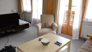 Ein Zimmer mit Fernseher, einer Couch, einem Sessel, einem Tisch mit Schale und Fernbedienung sowie einem Vorhangfenster.