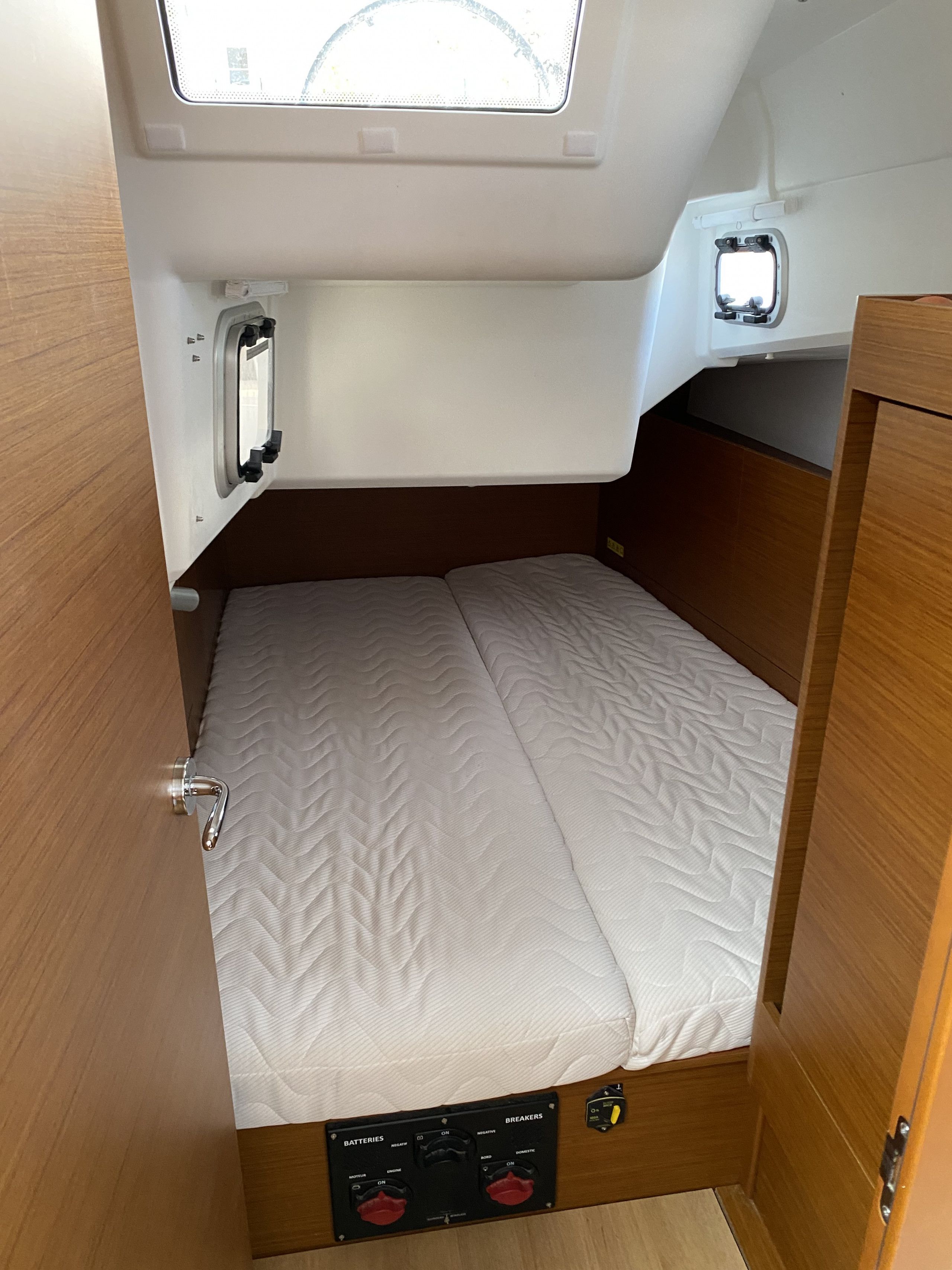 Interior compacto de una caravana con cama, panel de control para baterías y disyuntores, y ventana que proporciona luz natural.