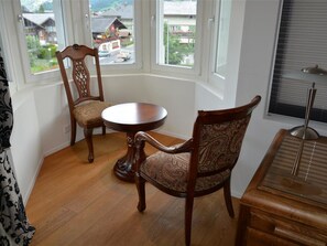 A corner room with a piano, two chairs, a round table, and a view of a street through large windows. - Haus am Bach Appartement/Fewo, Dusche oder Bad, WC 8-Bettwohnung by Interhome (Zweisimmen)