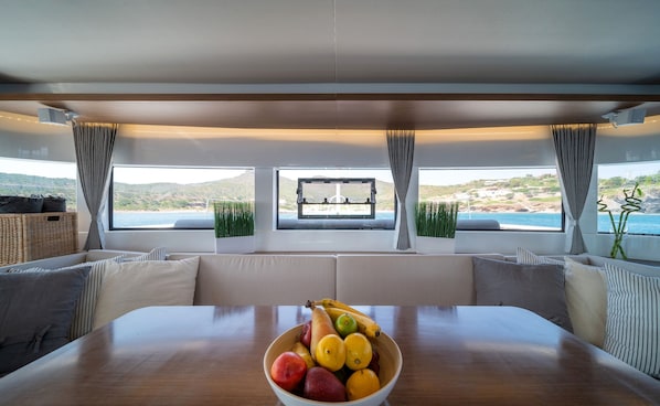 A dining area with a table and a bowl of fruit, large windows with curtains, and a view of the sea and hills.
