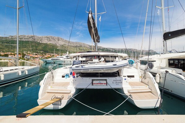 Marina con un catamarán blanco llamado 'Madame El Grande Split' amarrado, rodeado de otras embarcaciones y con montañas al fondo.