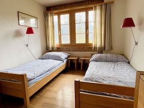 A room with two wooden beds, a small table, and a window with curtains. - Akelei (gross) Appartement/Fewo, Dusche oder Bad, WC 7-Bettwohnung by Interhome (Zweisimmen)