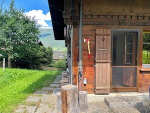 A rustic wooden building with a wooden door, a window, and a view of a green landscape with trees and mountains in the background. - Akelei (gross) Appartement/Fewo, Dusche oder Bad, WC 7-Bettwohnung by Interhome (Zweisimmen)