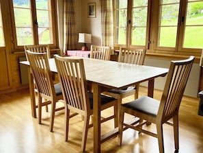 A dining room with a wooden table and chairs, large windows with curtains, and a view of greenery outside. - Akelei (gross) Appartement/Fewo, Dusche oder Bad, WC 7-Bettwohnung by Interhome (Zweisimmen)