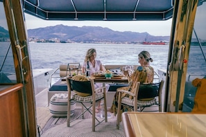 Uma área de refeições em um barco, com uma mesa posta para dois, cadeiras e vista para a água e montanhas.