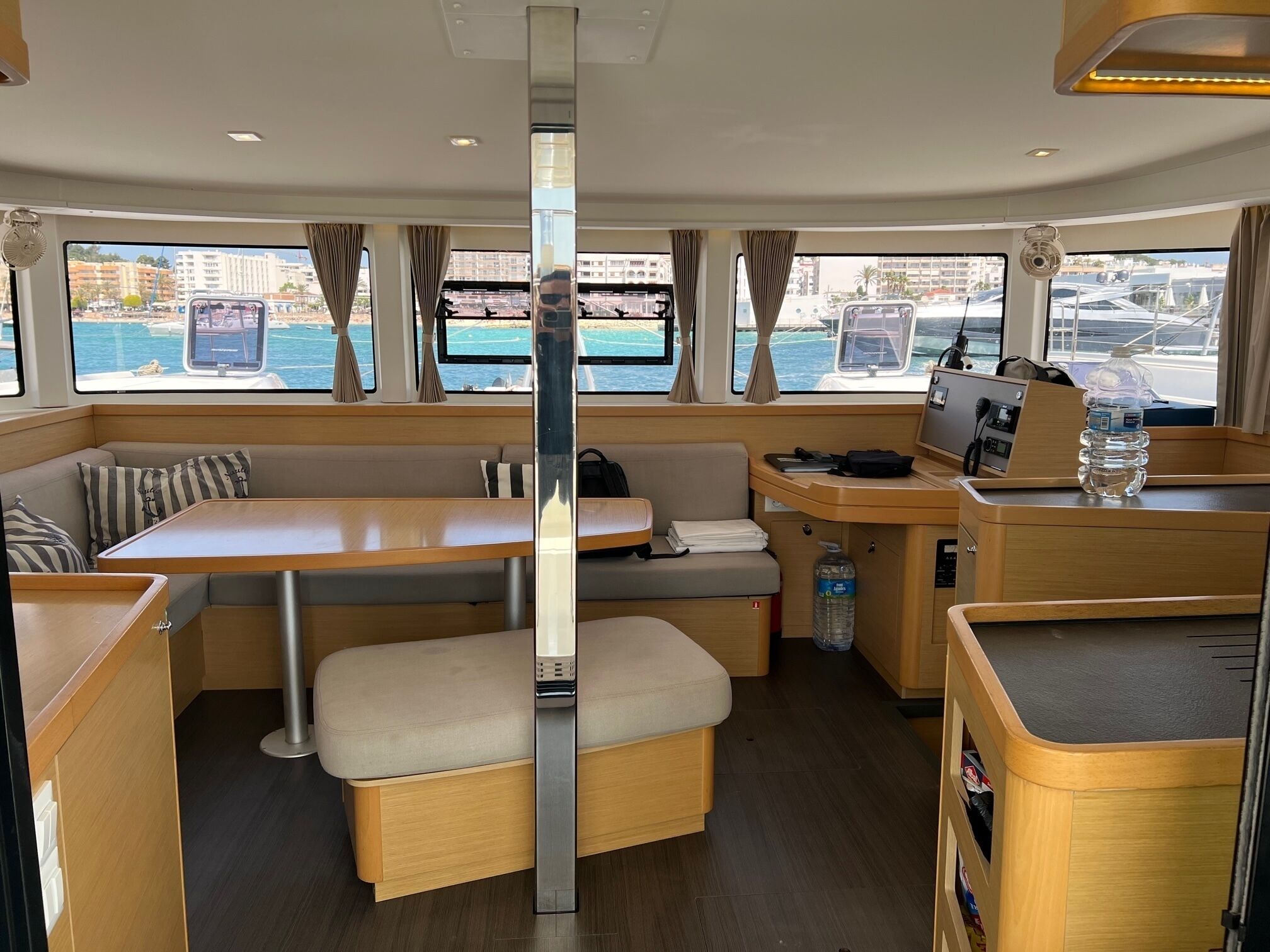 A compact interior with a central table, cushioned seating, a control panel, and a view of the marina through large windows.