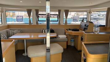 A compact interior with a central table, cushioned seating, a control panel, and a view of the marina through large windows.