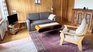 Ein Wohnzimmer mit grauem Sofa, einem Holzschrank, einem kleinen Tisch mit Tastatur, einem Fernseher auf einem Holzgestell und einem gemusterten Teppich.