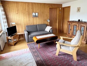 Ett vardagsrum med en grå soffa, ett träskåp, ett litet bord med ett tangentbord, en tv på ett träställ och en mönstrad matta.