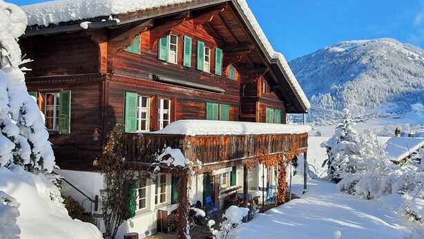 Eine Holzhütte mit schneebedecktem Dach, grünen Fensterläden und Bergelandschaft.