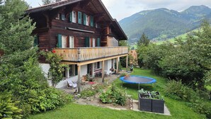 Eine Holzchalet mit einem Balkon, einem Garten inklusive Trampolin und einer Bergkulisse.