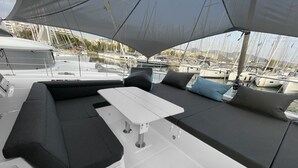 A yacht deck with a white table, black and grey cushioned seating, and a view of the marina in the background. - Sun-Kissed Sailing Adventure in Islas Sarónicas (Alimos)