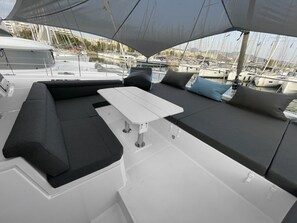 A yacht deck with a white table, black and grey cushioned seating, and a view of the marina in the background. - Sun-Kissed Sailing Adventure in Islas Sarónicas (Alimos)