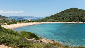 A coastal landscape with clear blue waters, a sandy beach, and a hill with dense vegetation.