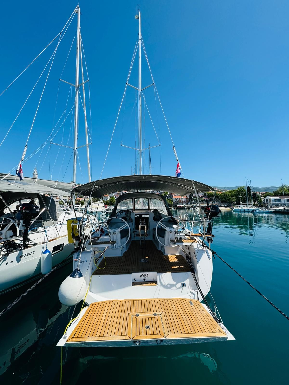 A sailboat docked at a marina with a wooden deck, multiple masts, and a clear blue sky.
