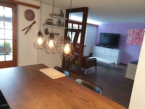 A dining area with a wooden table, hanging light fixtures, and a view into a living room with a television and shelves.