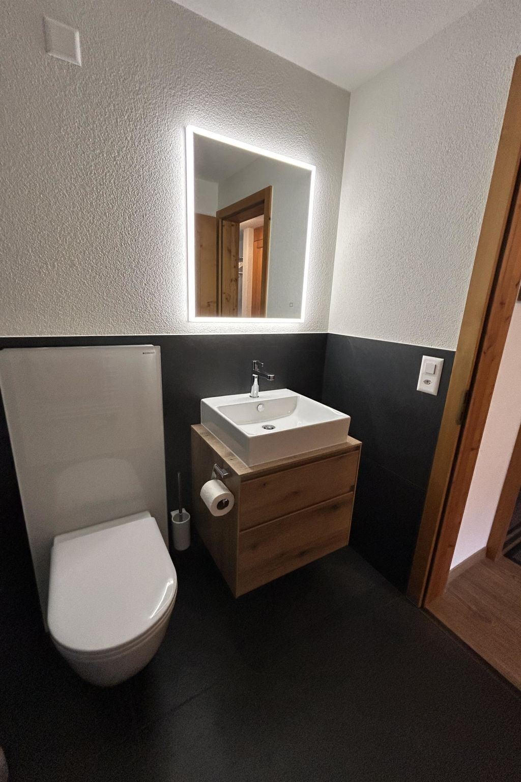 Ein kleines Badezimmer mit Toilette in Weiß, einem Holzwaschtisch mit weißem Waschbecken und einem Spiegel mit integrierter Beleuchtung.