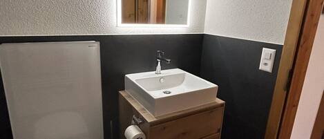 Ein kleines Badezimmer mit Toilette in Weiß, einem Holzwaschtisch mit weißem Waschbecken und einem Spiegel mit integrierter Beleuchtung.