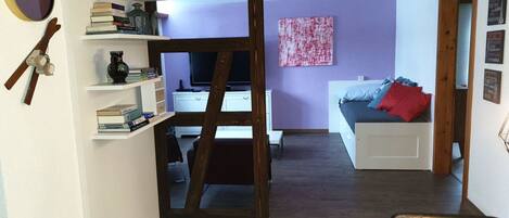 A room with a desk, shelves with books, a ladder, a bed with pillows, and a wall with a painting.