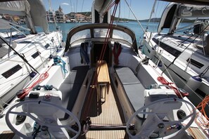 Ein Segeldeck mit mehreren Masten, einem Steuerrad und Blick auf einen Yachthafen mit Gebäuden im Hintergrund.