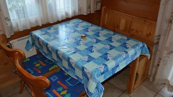 Une table à manger recouverte d’une nappe bleue à motifs, des chaises en bois avec des coussins bleus, et une fenêtre ornée de rideaux légers.