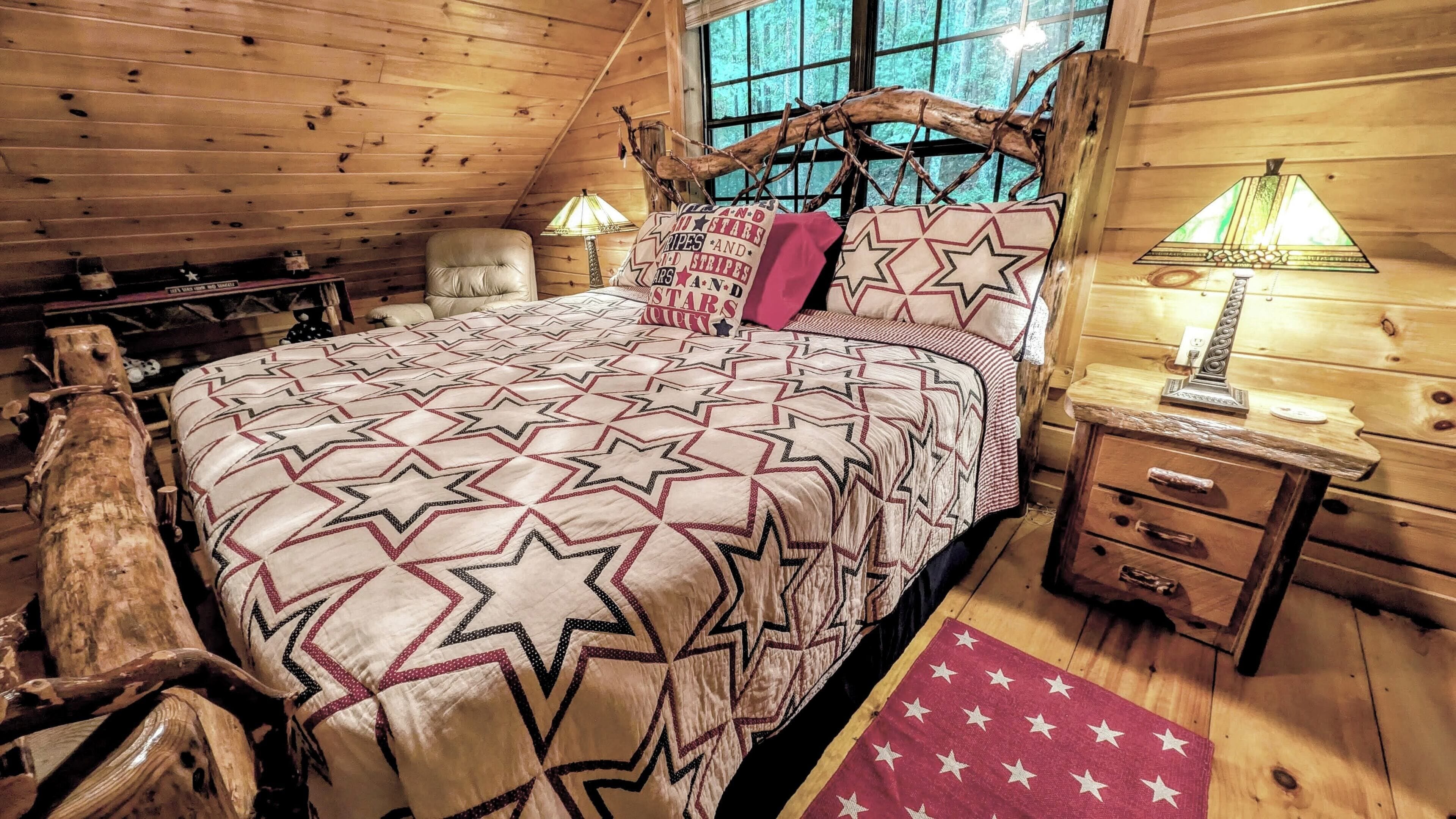 A log cabin bedroom with a star-patterned bedspread, a wooden headboard, a nightstand with a lamp, and a view of the forest through the window.