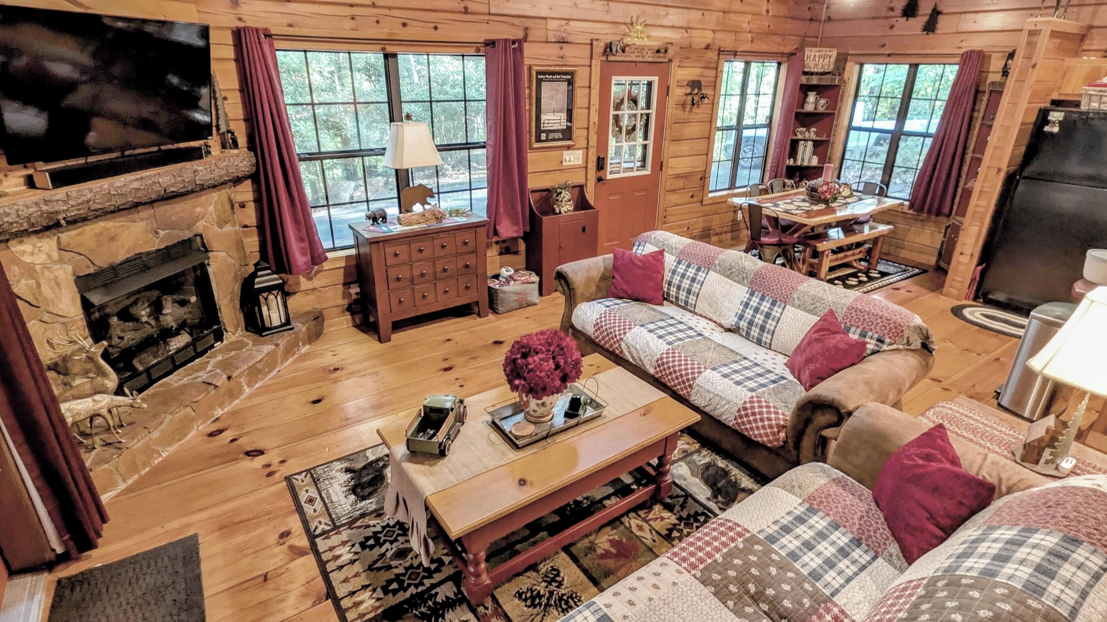 A cozy cabin interior with a stone fireplace, wooden furniture, and large windows.