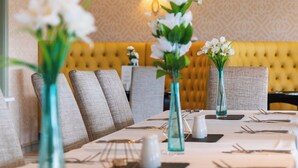 Un espace repas avec des tables dressées, des vases remplis de fleurs et, au fond, une tête de lit capitonnée.