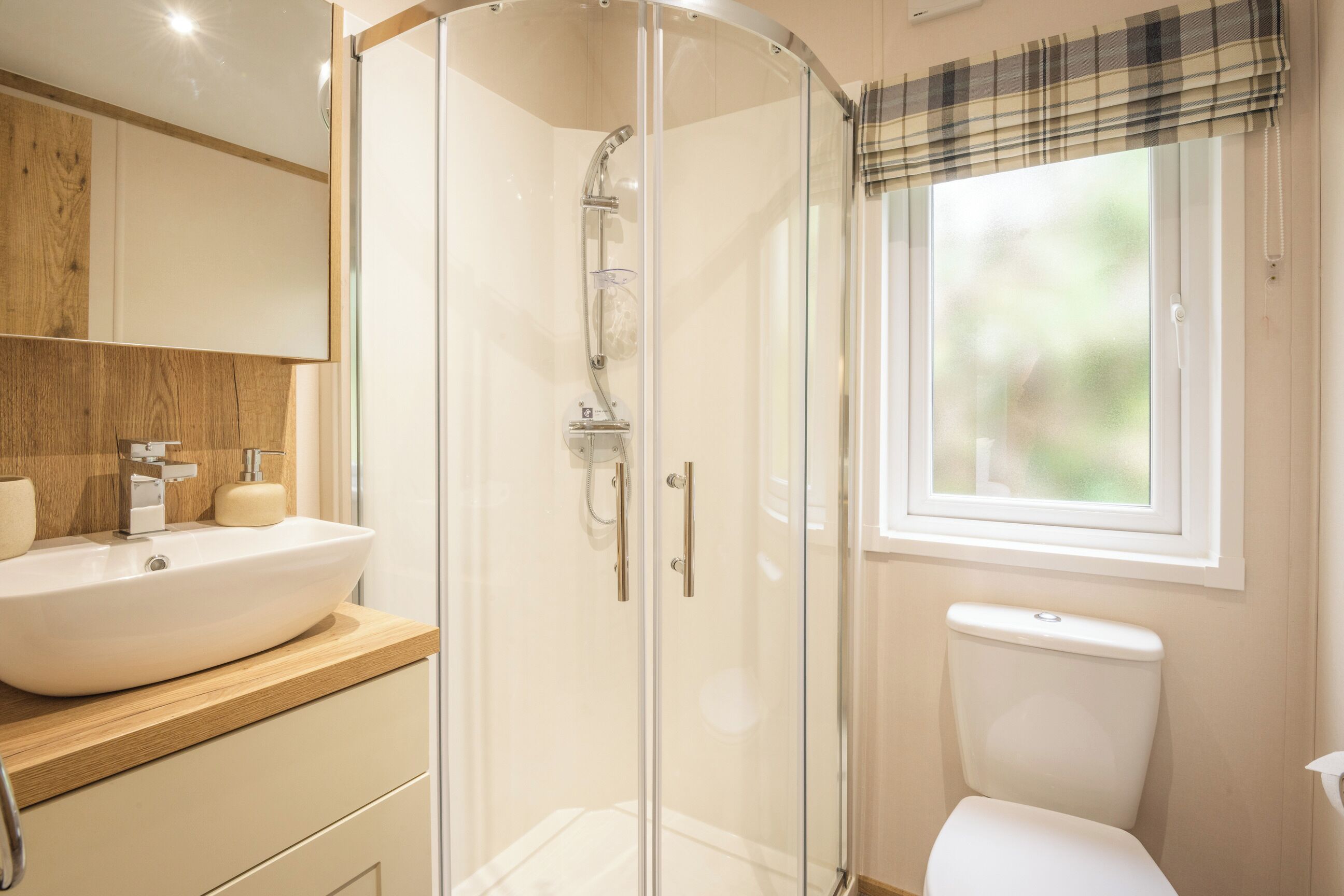Un bagno con doccia vetrata, un lavandino bianco e una finestra con tenda a quadri.