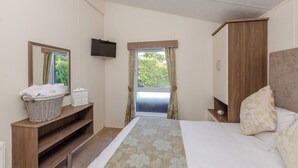 Une chambre à coucher comprenant un lit, une télévision, une fenêtre avec des rideaux, une armoire en bois et une petite étagère avec un panier et un récipient.