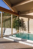 Piscina interior moderna con paredes de vidrio transparente, techo de madera y vistas al exterior a zonas verdes.