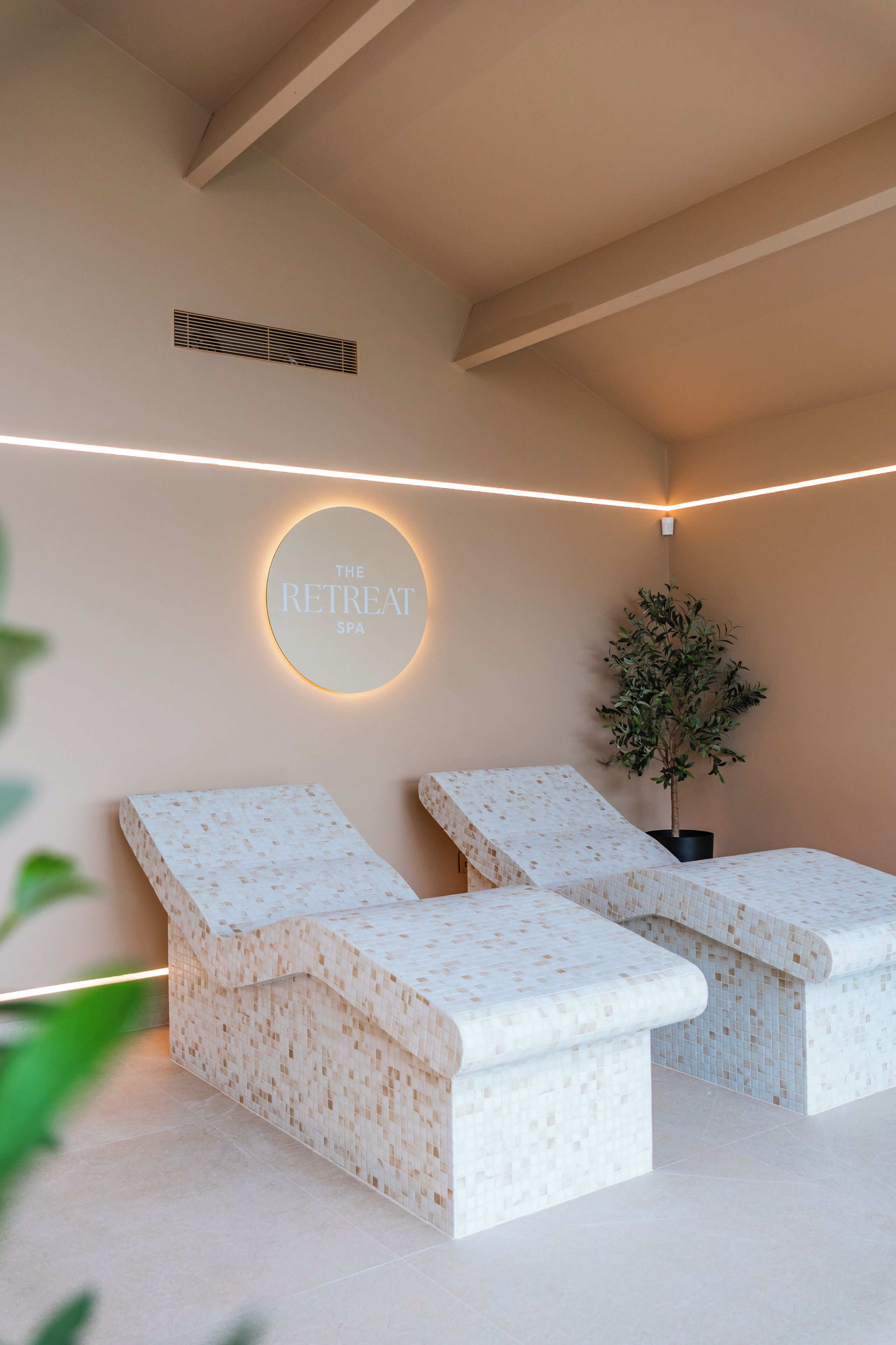 Ein Spa-Raum mit zwei Massagetischen, einem an der Wand befestigten Schild mit der Aufschrift 'The Retreat Spa' und einer Zimmerpflanze.