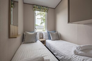 A bedroom with two single beds, a window with a patterned curtain, a wooden shelf, and a mirror.