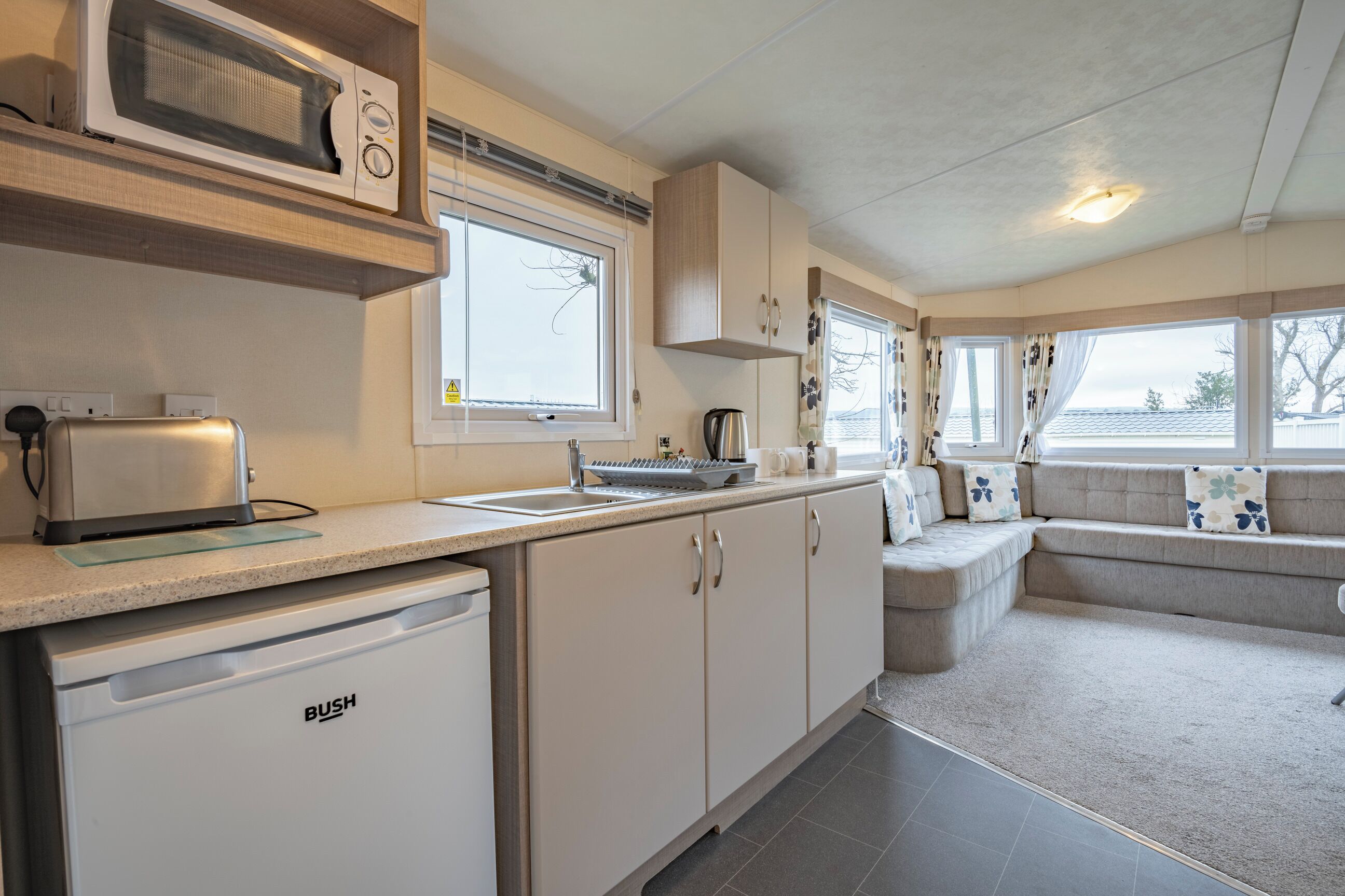 A compact kitchen with a microwave, toaster, and dishwasher. There is a seating area with a cushioned bench and a window with curtains.
