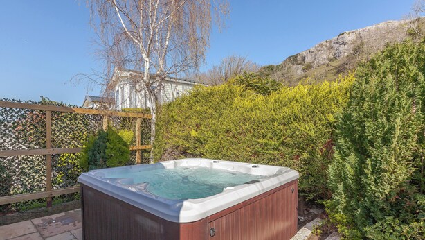 Un bain à remous installé à l’extérieur, sur une terrasse en bois, entouré de verdure et avec une maison en arrière-plan.