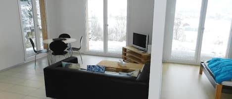 A spacious living room with a black sofa, a wooden coffee table, and a television on a wooden stand. There are two windows providing a view of the outside.