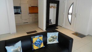 Un salón con un sofá negro, dos cojines, una cocina al fondo y una puerta con ventanal.