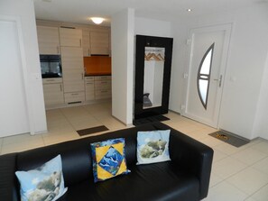 A living room with a black sofa, two cushions, a kitchen in the background, and a door with a window. - Schön-Egg Appartement/Fewo, Dusche, WC 2-Bettwohnung by Interhome (Zweisimmen)