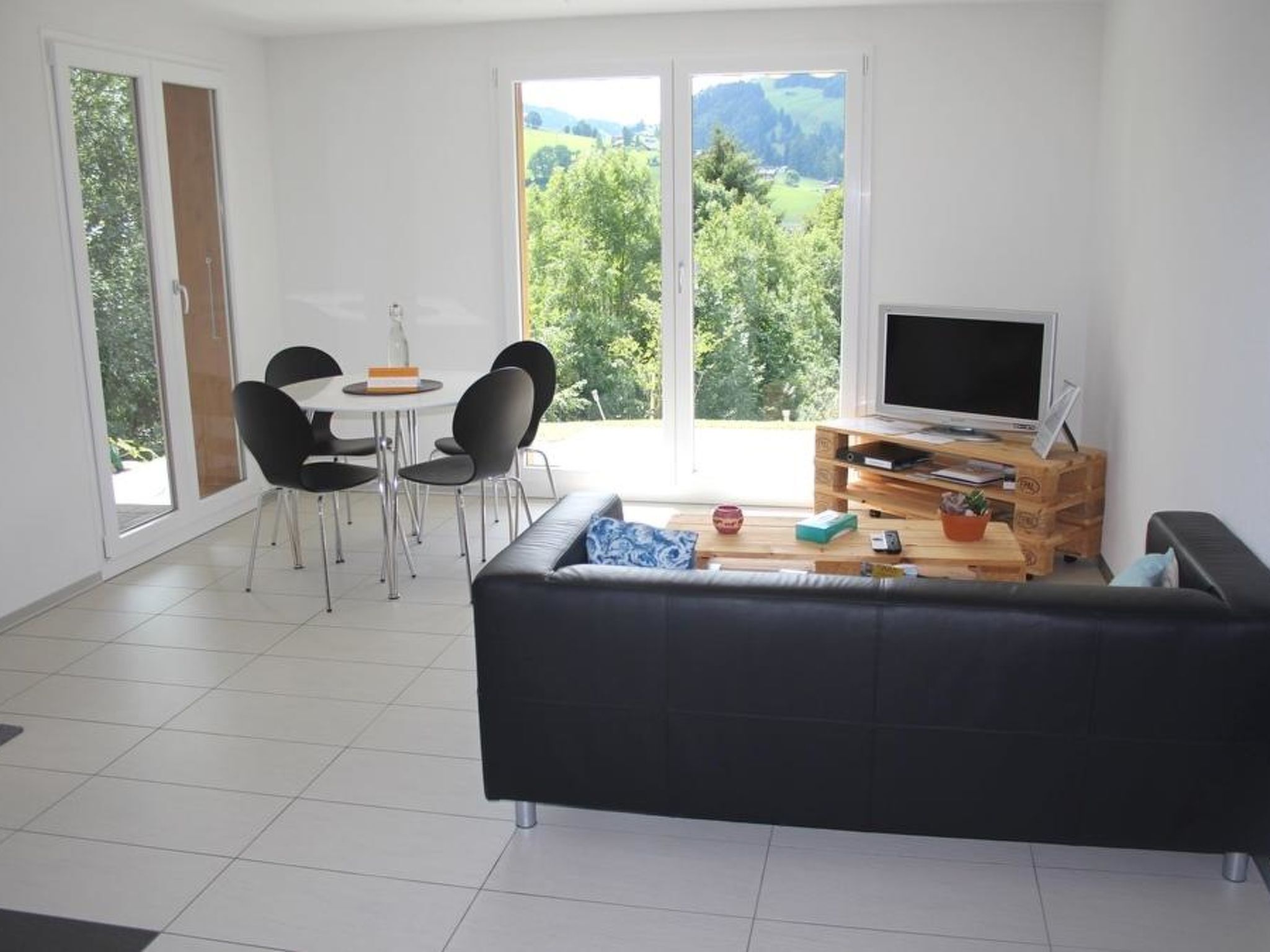 A modern living room with a black sofa, a dining table with chairs, a TV on a wooden stand, and large windows with a view of greenery.